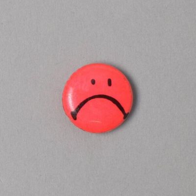 Miniature circular pin badge with a frowning "Smiley Face" printed on fluorescent red ground; laminated paper, cardboard, metal, by Button Up, New York, 1980s